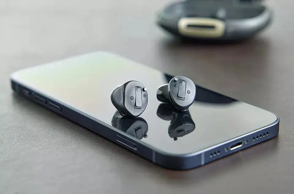 Two discreet in-ear hearing aids displayed beside a smartphone and charging case